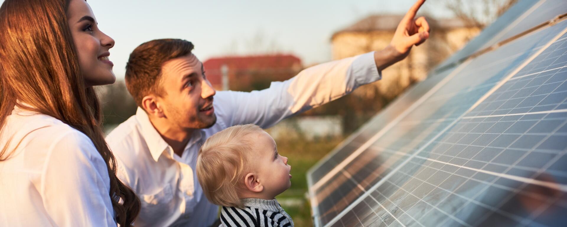 Mann steht mit seiner Frau und einem Kind vor einer Solaranlage und zeigt in Richtung Sonne
